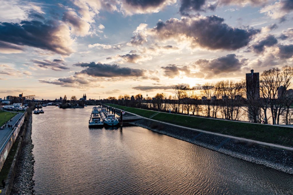 Blick auf den Rhein am Mülheimer Hafen. Im Hintergrund geht die Sonne am bewölkten Himmel unter