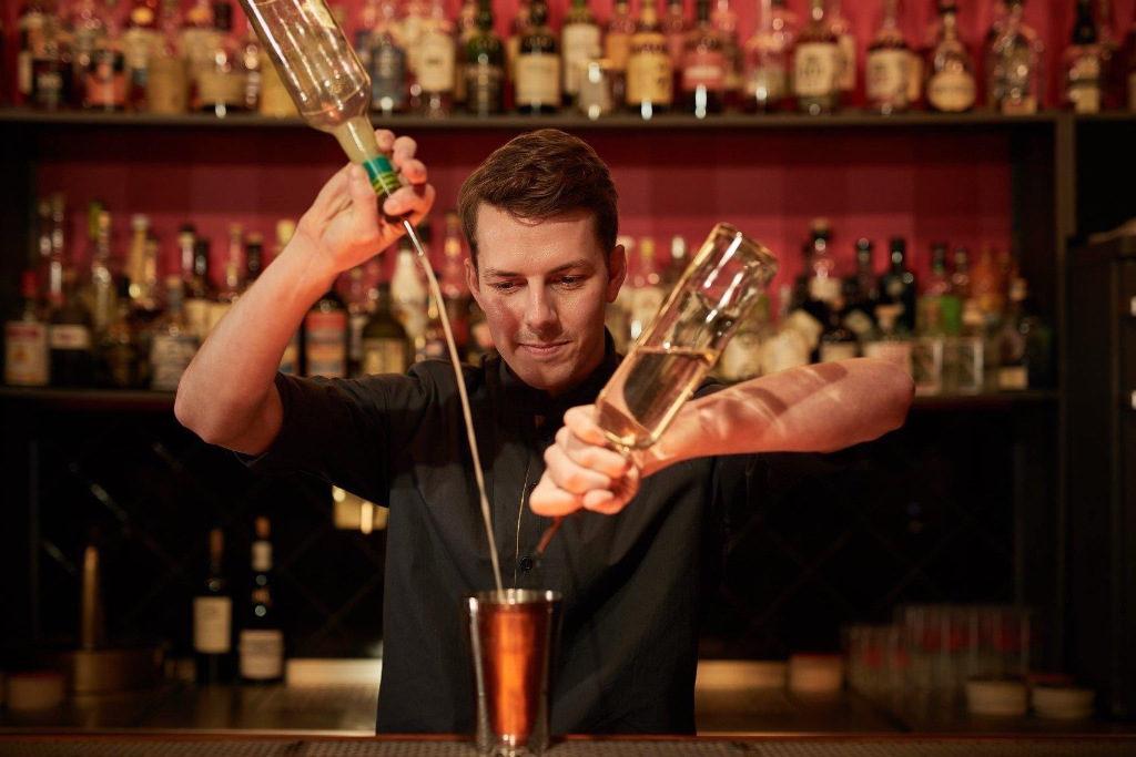 Barkeeper mit zwei Flaschen in der Hand mixt einen Cocktail an einer Bar.