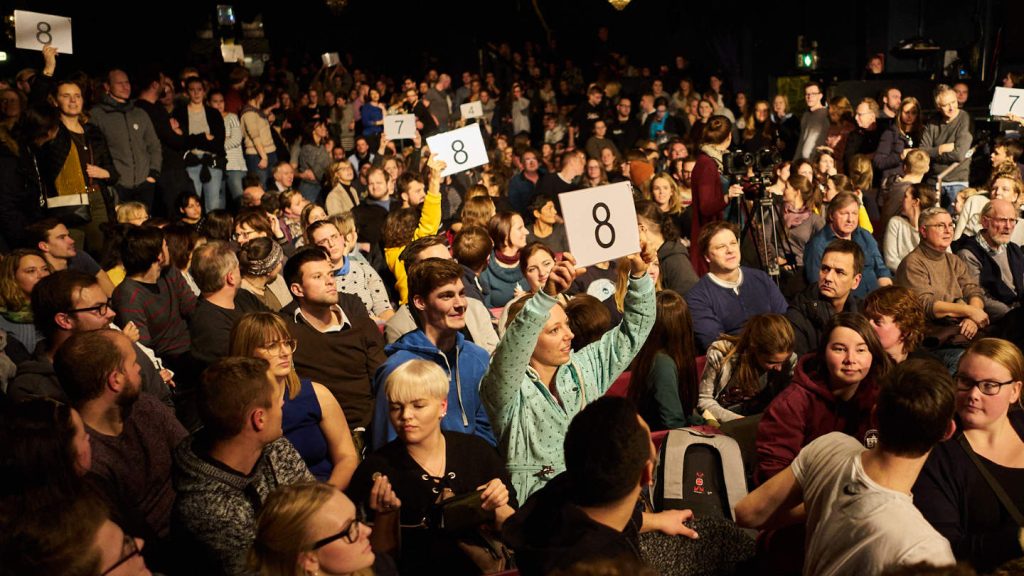 Aufnahme aus dem Publikum beim Science Slam. Menschen halten Zettel mit Zahlen in die Luft