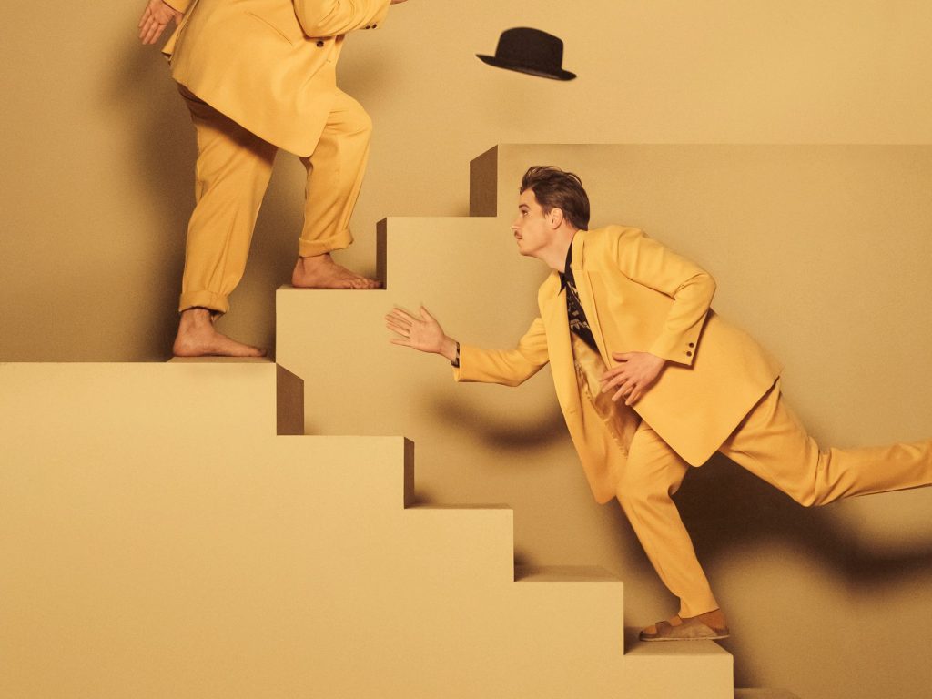 Zwei Männer in sandgelber Kleidung gehen eine sandgelbe Treppe hoch. Die Wand hinter ihnen ist ebenfalls sandgelb.