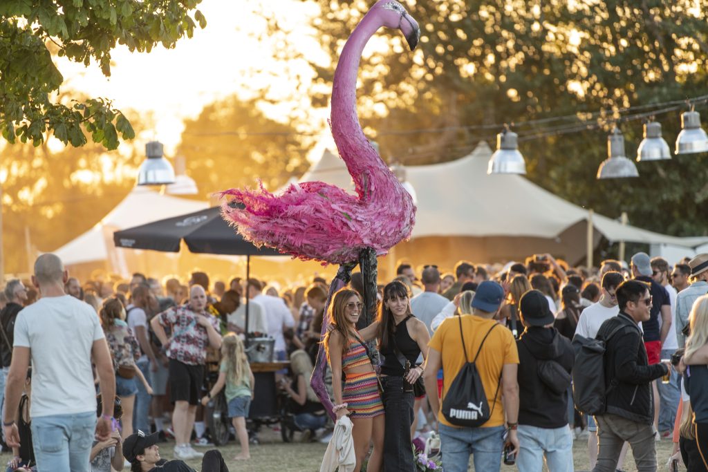 Auf dem Festivalgelände haben sich viele Menschen versammelt. Viele von ihnen tanzen. Die Sonne geht gerade unter. Im Bildmittelpunkt stehen zwei junge Frauen vor einer großen Nachbildung eines Flamingos und lassen sich fotografieren.