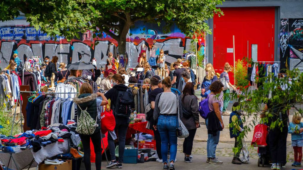 Menschen stehen an Flohmarktständen. Im Hintergrund eine Wand mit Graffiti und eine rote Tür.