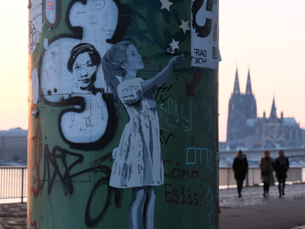 Paste-up eines Mädchen auf einer grünen Säule. Im Hintergrund der Kölner Dom im Sonnenuntergang.