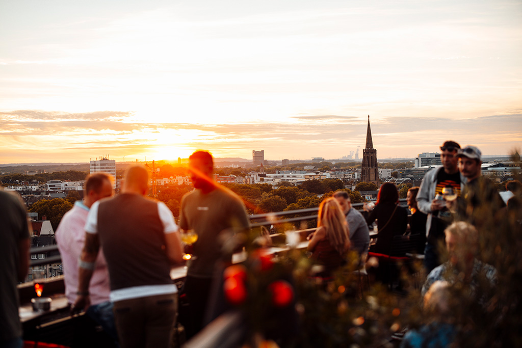 Eine Aufnahme von der Dachterrasse der Bar Botanik, die Sonnenuntergangsstimmung zeigt.  Leute die Cocktails trinken und sich unterhalten. Im Hintergrund die Skyline von Köln.