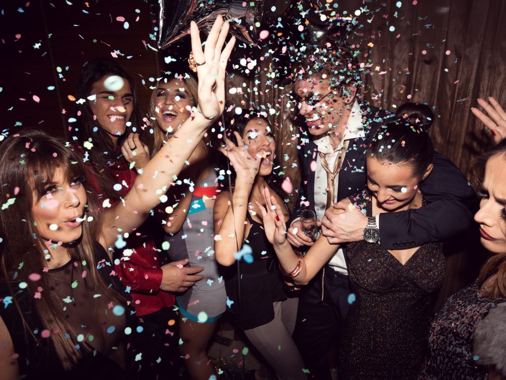Eine Gruppe junger Menschen in Abendkleidung tanzt und feiert zusammen. Von oben rieselt buntes Konfetti.