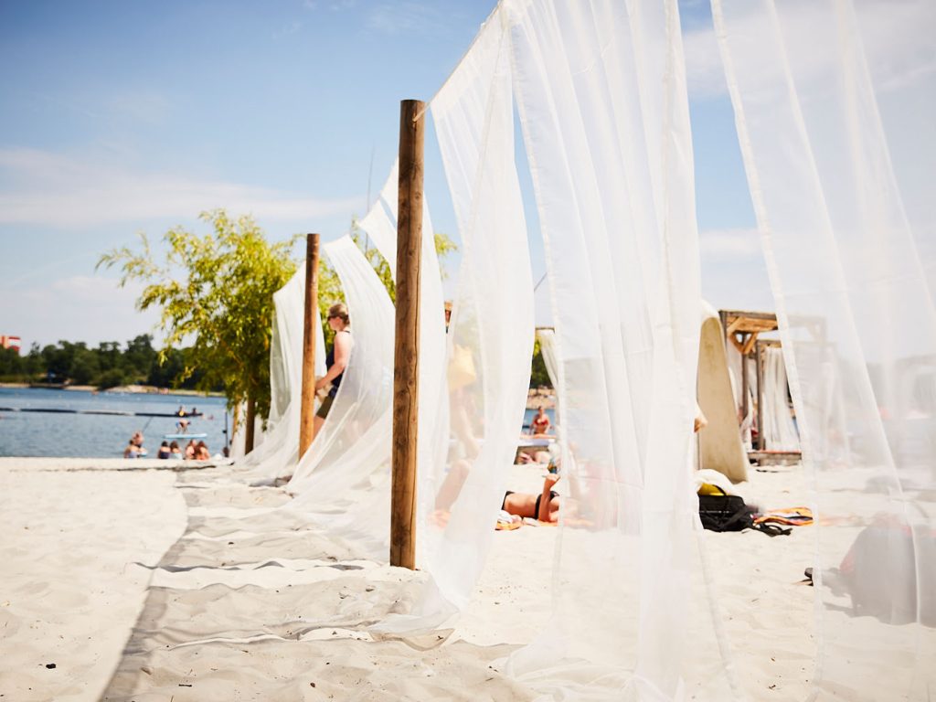 Beach Clubs Köln: Dieser Beach Club beeindruckt mit weißem Sandstrand, See und weißen Himmelbetten.