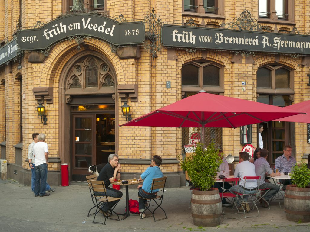 Brauhäuser in Köln: Ein ziegelsteiniges Brauhaus von außen, vor dem Gäste an Tischen Bier trinken und sich unterhalten.