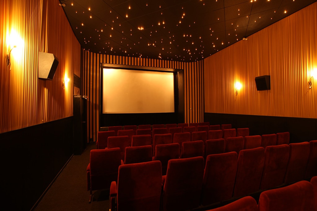 Man sieht einen alten Kinosaal mit roten Samtsitzen und einer kleinen Leinwand. 