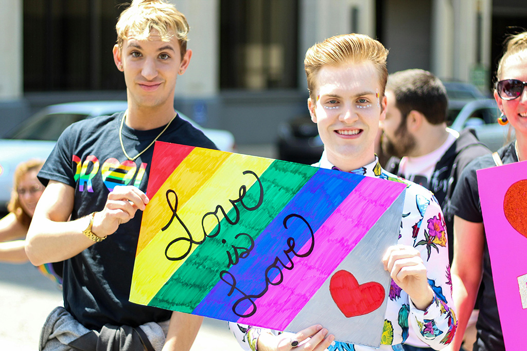 Zwei junge Männer halten ein buntes Schild in die Höhe auf dem "Love is Love" geschrieben steht. 