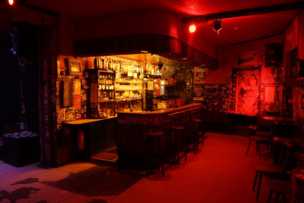 Das Sonic Ballroom ist in rotes Licht getaucht. Die Bar ist erleuchtet, am Tresen stehen leere Barhocker. Die Wönre sind mit Postern und Stickern beklebt. Foto: Dirk Heinrich