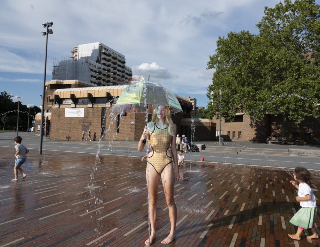 Das Model Olga läuft an einem heissen Sommertag in einem neongrünen Badeanzug über einen Platz mit Brunnen in Köln Chorweiler. In der Hand hält sie einen Regenschirm. Foto: Anne Schönharting