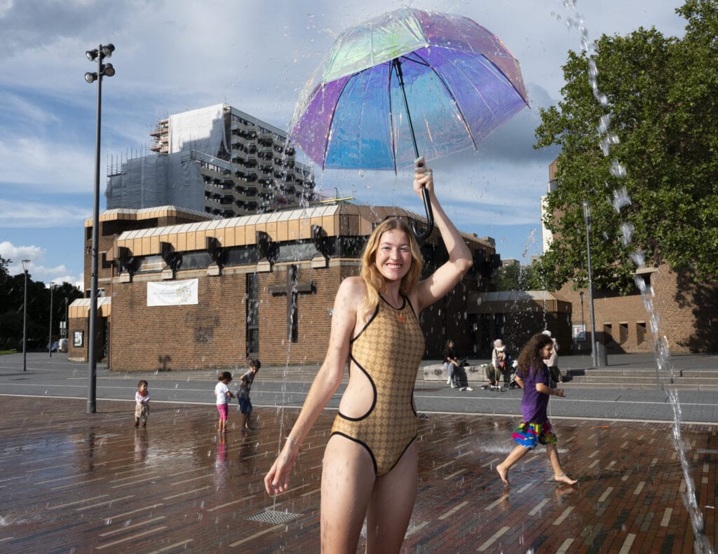 Das Model Olga läuft an einem heissen Sommertag in einem neongrünen Badeanzug über einen Platz mit Brunnen in Köln Chorweiler. In der Hand hält sie einen Regenschirm. Foto: Anne Schönharting